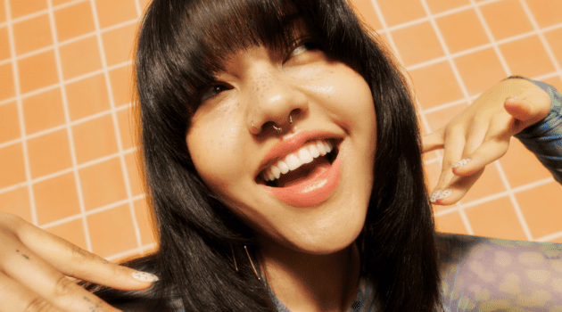 Smiling person with straight bangs and septum piercing, showcasing trendy nails against a tiled background.