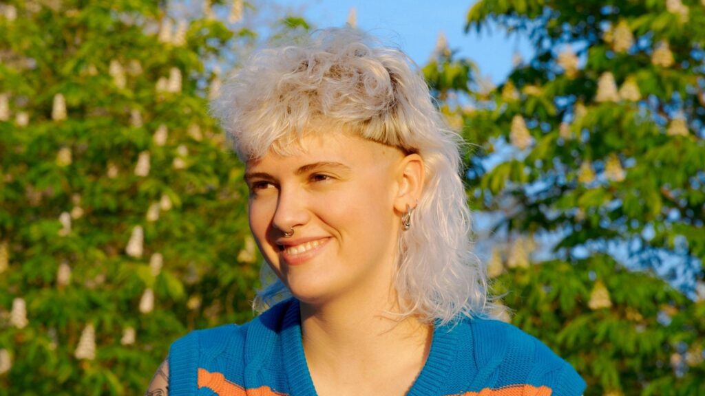 [ALT_TEXT: A young woman with a curly white blonde mullet haircut sitting in a park on a sunny day.]