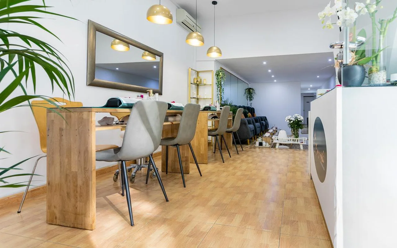 A bright nail salon featuring long wooden manicure tables with grey modern chairs. Gold dome pendant lights hang above the stations, and a large mirror with a dark frame is mounted on the white wall.