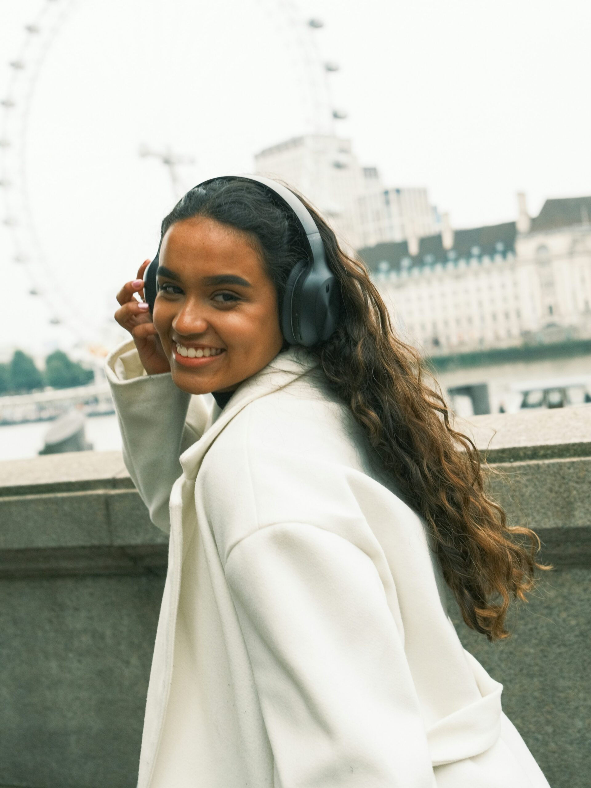 A smiling woman with microblading on her brows wearing black over-ear headphones and a white coat walking along a canal in Amsterdam