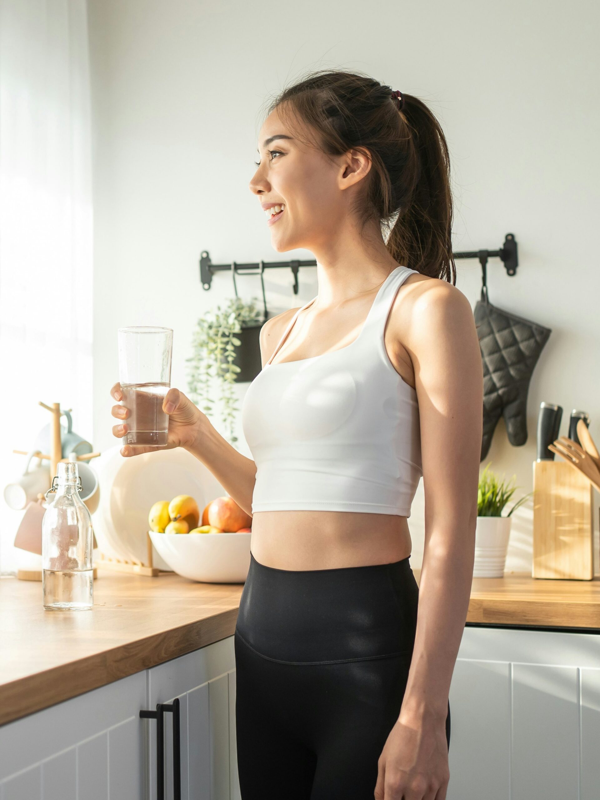 Side view of a woman drinking water in her kitchen in front of a sunny window