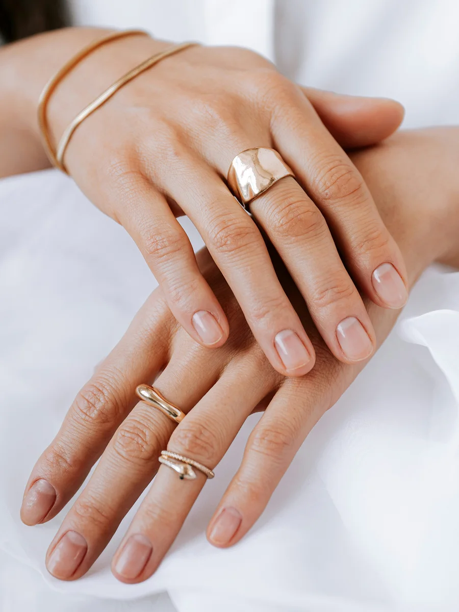 Close-up of two hands resting on a white sheet showing healthy, natural nails and adorned with rose-gold rings and bracelets