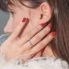 Close-up side view of a brunette woman’s head, her hand touching her face showing a glossy red gel manicure