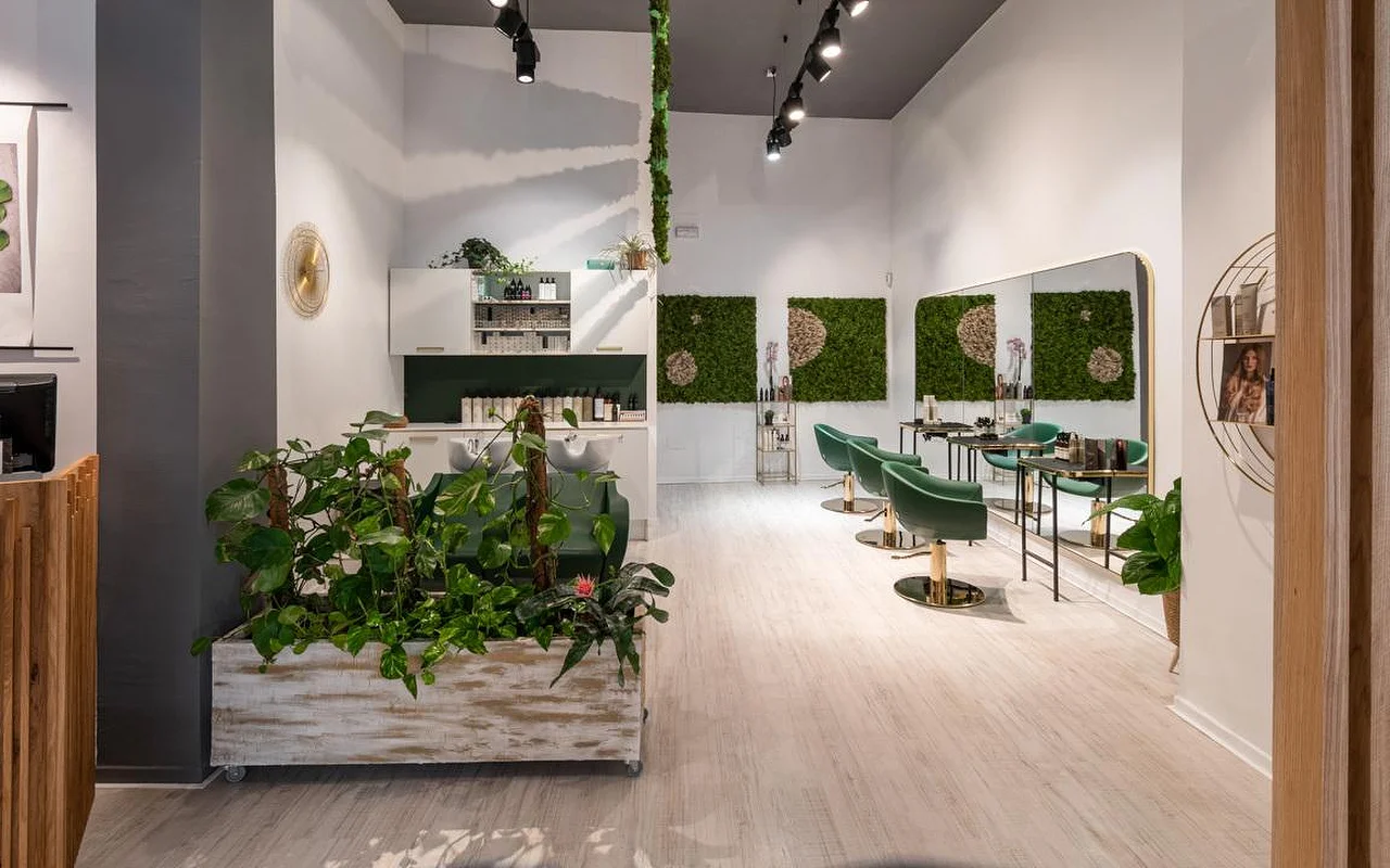 A plant-filled hair salon with pale wood floors, an aged white planter overflowing with greenery, green moss wall art, emerald-green velvet styling chairs with gold detailing, and a wash station behind.