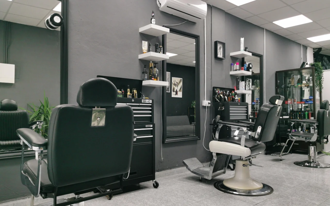A traditional barbershop with charcoal-grey walls, classic black leather barber chairs with chrome bases and footrests, white floating shelves stocked with grooming products, and large mirrors on tiled flooring.